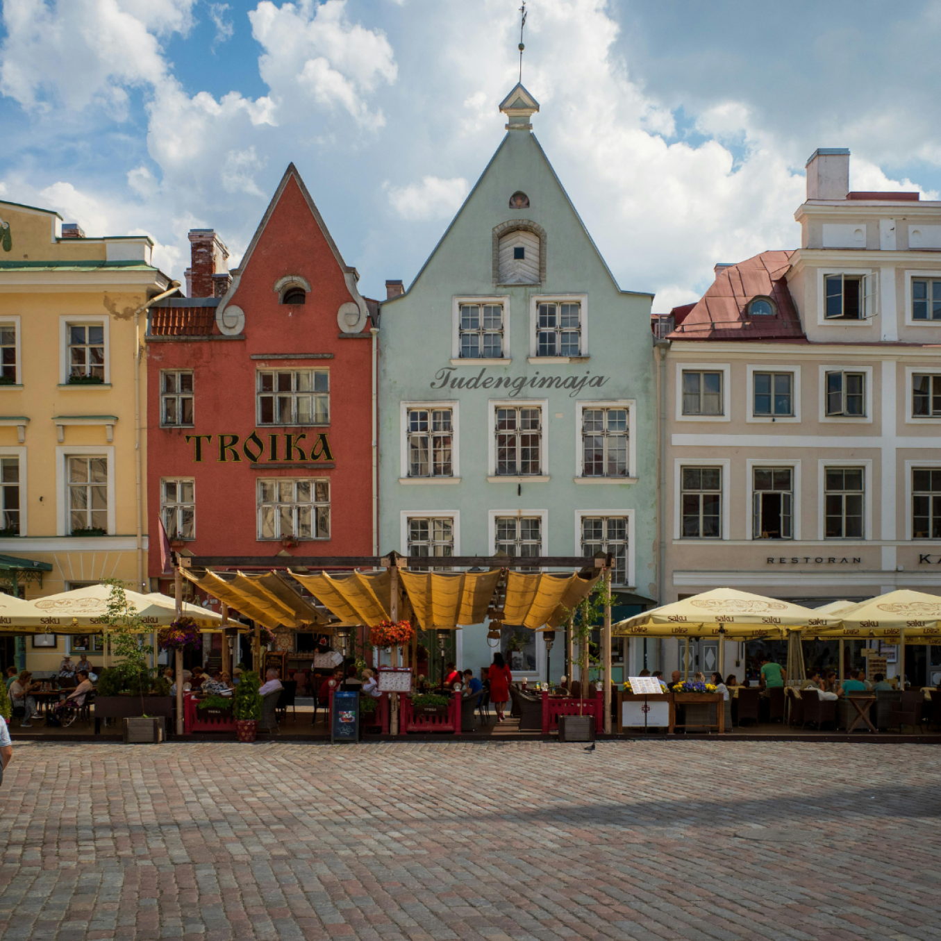 Värikkäitä, kapeita vanhoja kaupunkitaloja mukulakivitorin laidalla. Rakennusten julkisivut ovat pastellinsävyisiä – keltainen, punainen ja vaaleanvihreä – ja niissä on koristeelliset päädyt. Katutasossa on ravintoloita terasseineen, aurinkovarjoineen ja markiiseineen, ja ihmisiä istuu ulkopöydissä kesäisen pilvitaivaan alla.